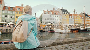 A woman sits on the embankment and admires the colorful buildings on the banks of the Nyhavn canal in Copenhagen