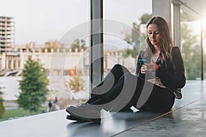 Woman sits on floor near window, uses smartphone.Hipster girl looking on screen of phone,blogging, browsing internet.