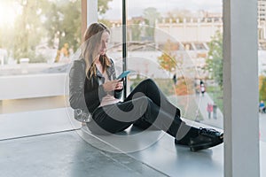 Woman sits on floor near window, uses smartphone.Hipster girl looking on screen of phone,blogging, browsing internet.