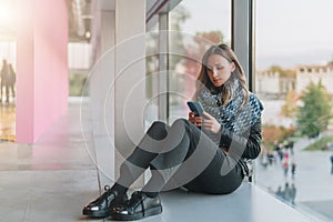 Woman sits on floor near window, uses smartphone.Hipster girl looking on screen of phone,blogging, browsing internet.