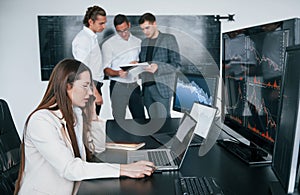 Woman sits in front of laptop and displays. Team of stockbrokers works in modern office with many display screens