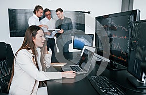 Woman sits in front of laptop and displays. Team of stockbrokers works in modern office with many display screens