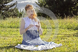 Woman sits grass in the park and knits wool clothes on knitting needles.