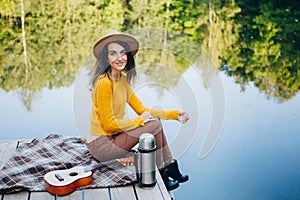 Woman sits with a guitar on a bridge on a lake with an autumn landscape