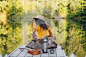 Woman sits with a guitar on a bridge on a lake with an autumn landscape