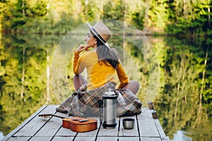 Woman sits with a guitar on a bridge on a lake with an autumn landscape