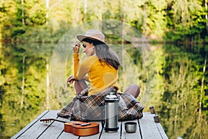 Woman sits with a guitar on a bridge on a lake with an autumn landscape