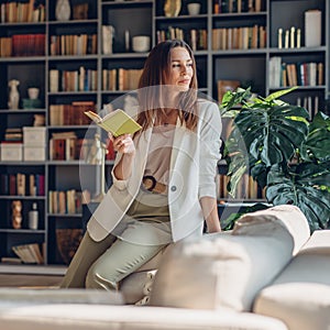 Woman sits at home with open book and looks thoughtfully away.