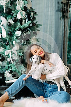 Woman sits near Christmas tree and plays with puppy of dalmatian dog