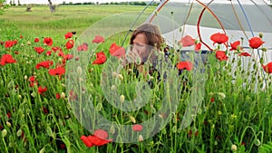 Woman sits near tent on blooming poppy field, admires and sniffs the flowers