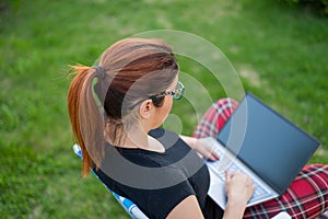 Woman sits in a park and types on the computer while shopping online. The girl maintains a social distance and works