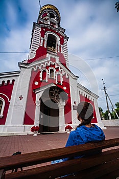 Woman sits on pew near to Church of the Presentation of the Lord in Dmitrov. View from behind on believing girl next to the red