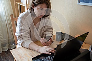 A woman sits with a phone in her hands and works at a laptop at home