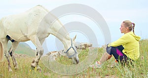 The woman sits, relax and looks at beautiful white horse. Love for pets. Mountain hill in steppe. Relaxing and