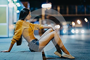 A woman sits on sidewalk against background of night city