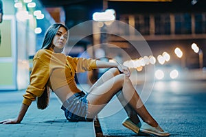 A woman sits on sidewalk against background of night city