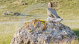 A woman sits on a stone in a lotus position and the wind blows her hair against the background of a feather grass