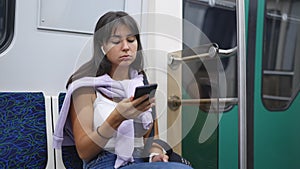 A woman sits in a subway car, puts on headphones, turns on music on her phone