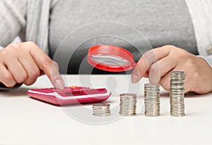 A woman sits at a table and counts on a calculator in the other hand a magnifying glass, a stack of coins on the table.