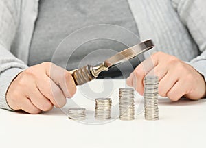 A woman sits at a table and holds a wooden magnifying glass over stacks of white coins. The concept of budget planning, savings.