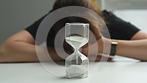 A woman sits at a table and looks at an hourglass, rubs her face with her hand in alarm. The woman is stressed that time