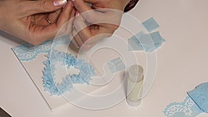 A woman sits at a table and makes a greeting card. Glues paper elements. Filmed from above