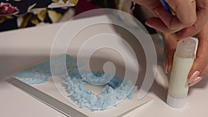 A woman sits at a table and makes a greeting card. Glues paper elements. Nearby on the table are tools and materials