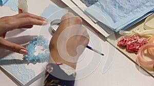A woman sits at a table and makes a greeting card. Glues paper elements. Nearby on the table are tools and materials. Filmed from