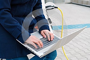 Woman sits on the trunk of a car and uses a notebook while the EV is charging