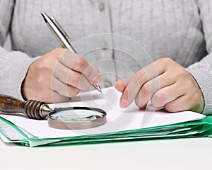 Woman sits at a white table and holds a metal pen over a pile of papers, in the other hand a magnifying glass