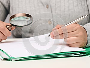 A woman sits at a white table and holds a metal pen over a pile of papers, in the other hand a magnifying glass. Finding mistakes