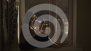 Woman sits on windowsill near wet window. Girl runs finger across glass.