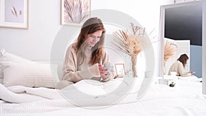 Woman sitting at bed at home, having breakfast and using smartphone