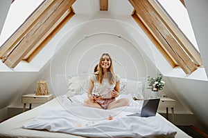 Woman is sitting on a bed with a laptop, use phone and drink coffee