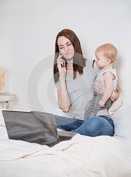 Woman sitting on a bed with a small child, working at a laptop and talking on the phone, a young mother working at home