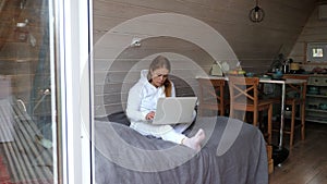 A woman is sitting on a bed in a triangular wooden house with a laptop.