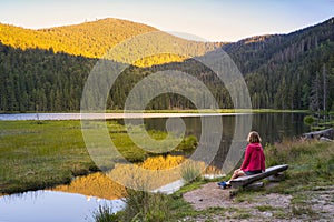 A woman sitting on a bench at beautiful Small Arber lake in the Bavarian Forest at sunset. View to mount GroÃŸer Arber