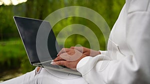 A woman is sitting on a bench and typing on a laptop. She is wearing a white shirt and white pants. Concept of