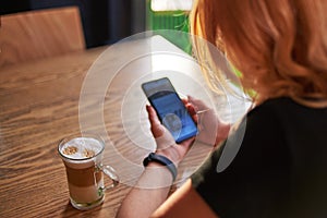 A woman is sitting in a cafe with a cup of cappuccino and is using a smartphone