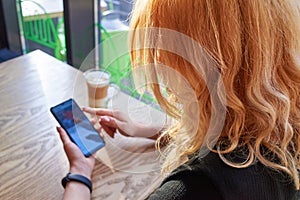 A woman is sitting in a cafe with a cup of cappuccino and is using a smartphone