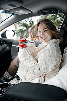 Woman sitting in a car in warm woolen sweater and socks holding cup of coffee