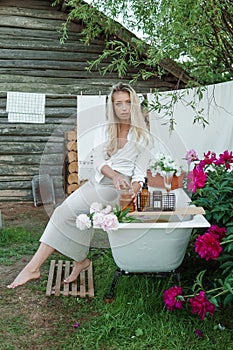 A woman is sitting on a cast-iron bathtub in the courtyard of a country house next to a bush of flowering peonies. The