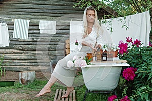 A woman is sitting on a cast-iron bathtub in the courtyard of a country house next to a bush of flowering peonies. The