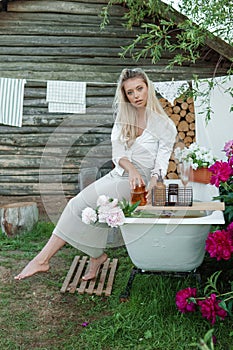 A woman is sitting on a cast-iron bathtub in the courtyard of a country house next to a bush of flowering peonies. The