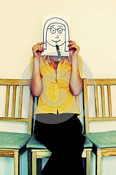 A woman sitting on a chair covering her face with a drawing of a girl with spectacles and blowout. Conceptual image shot