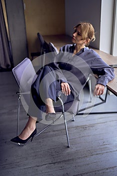Woman sitting on chair with legs crossed over the armrest. High heels, black pants, silk blouse. Cityscape outside through window