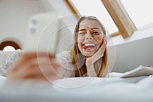 A woman is sitting comfortably on the bed, smiling and taking a selfie with her phone