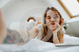 A woman is sitting comfortably on the bed, smiling and taking a selfie with her phone