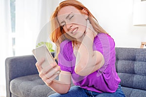 Woman sitting on couch holding phone suffers from neck pain