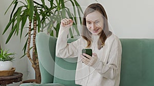 Woman sitting on the couch, using smartphone. Excited woman look at the screen, winning at the phone game